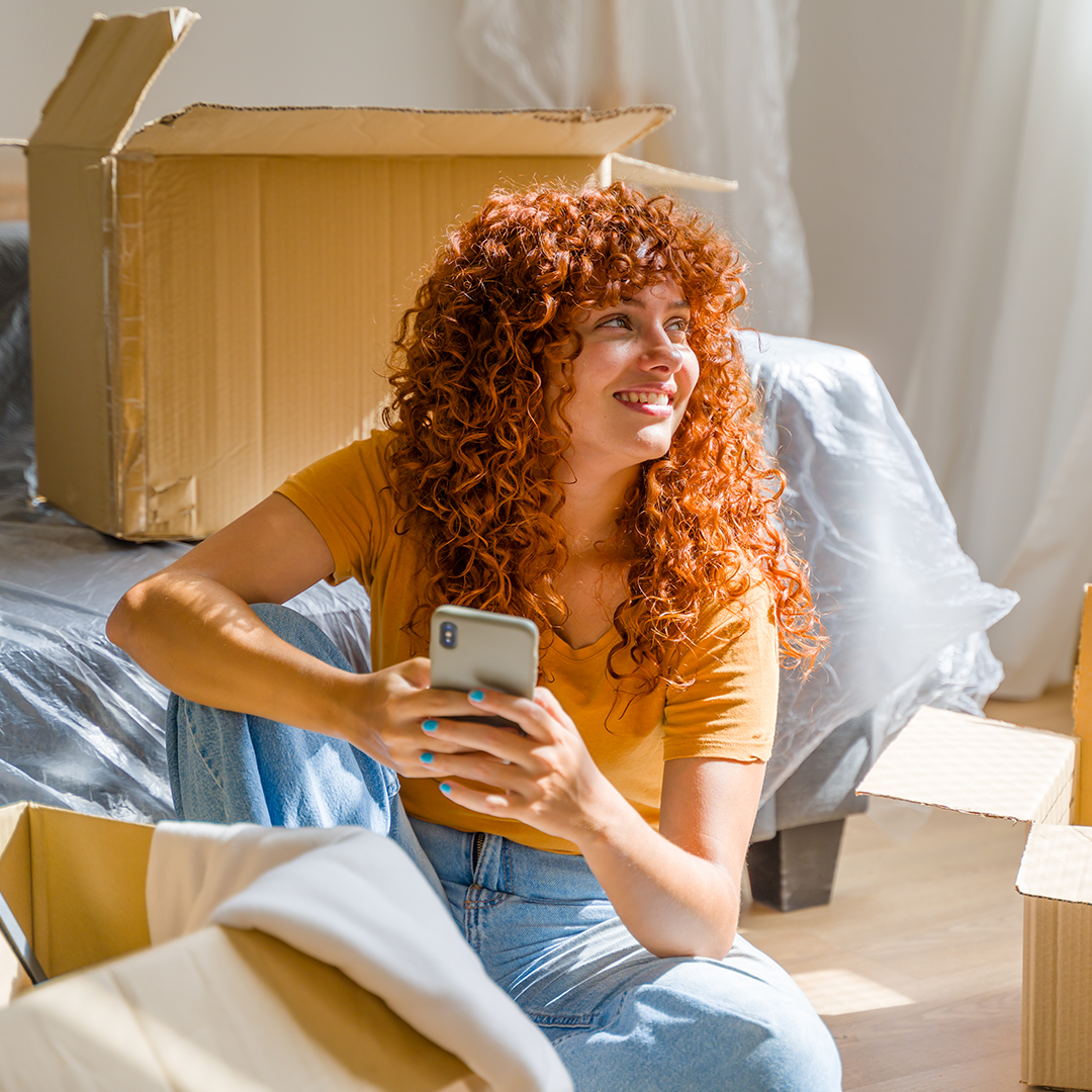 Image of a woman with curly red hair holding a cell phone contently smiling with moving boxes around her.