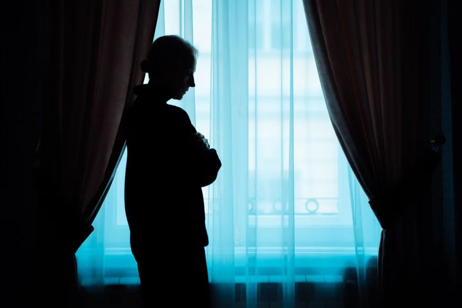 Person standing at a window in a dark room, looking lonely