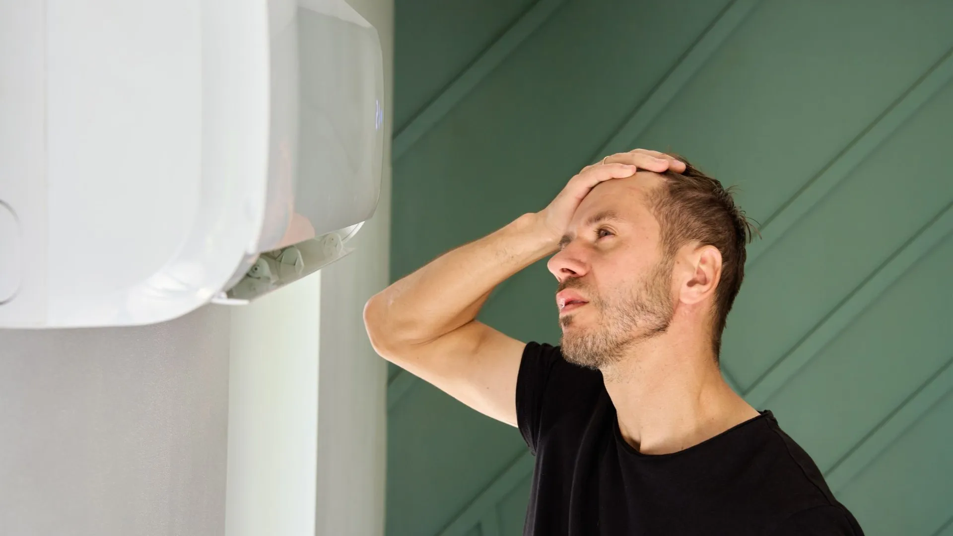 Man looking stressed and holding his forehead while standing next to a white air conditioning unit mounted on a wall.