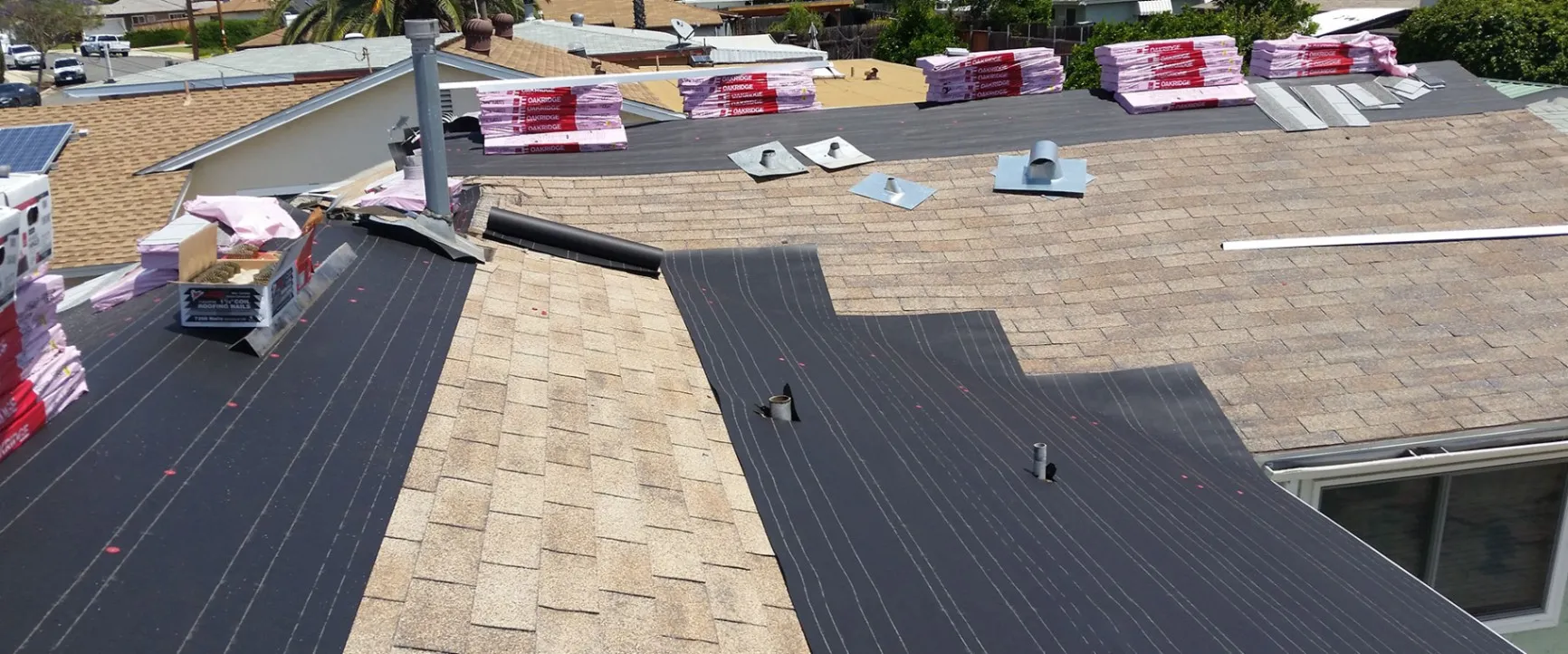 Roof installation in progress with exposed synthetic underlayment, vent flashing components, and bundles of asphalt shingles staged across the roof.