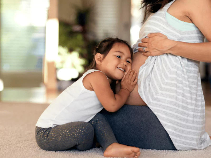 A young girl listens closely with her ear pressed against her mother's pregnant belly, sharing a quiet, intimate moment with her unborn sibling.