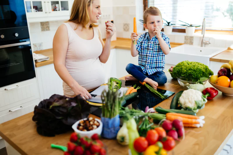 Pregnant mom snacking on vegetables with her young son on a kitchen counter, highlighting healthy Thanksgiving diet options for pregnant mothers.