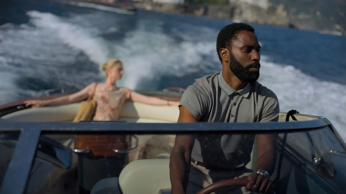 Two characters ride a speedboat on bright blue Mediterranean water, a coastal cliff visible in the distance behind them.