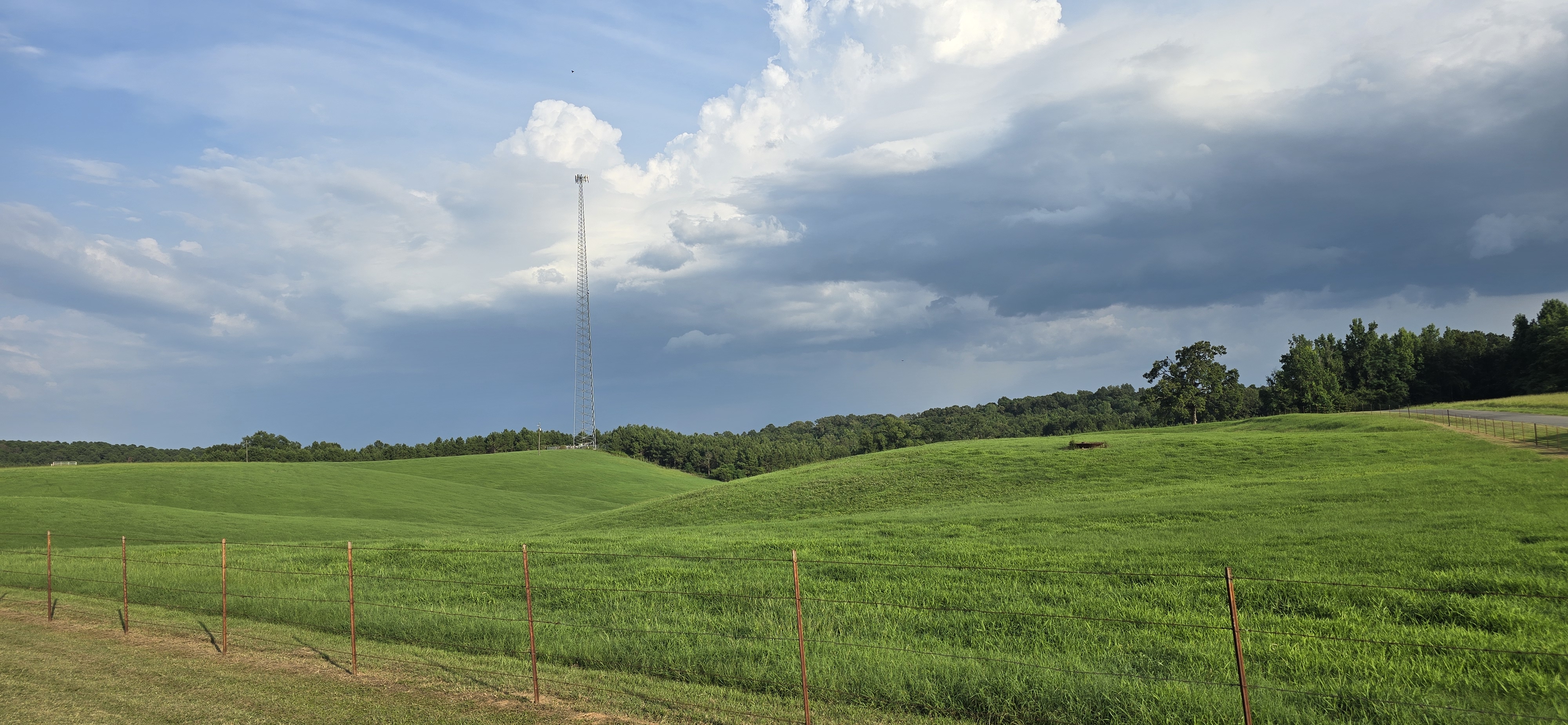 Cell tower in the rolling hills of North Louisiana surrounded by green grass and a beautiful cloud covered sky.