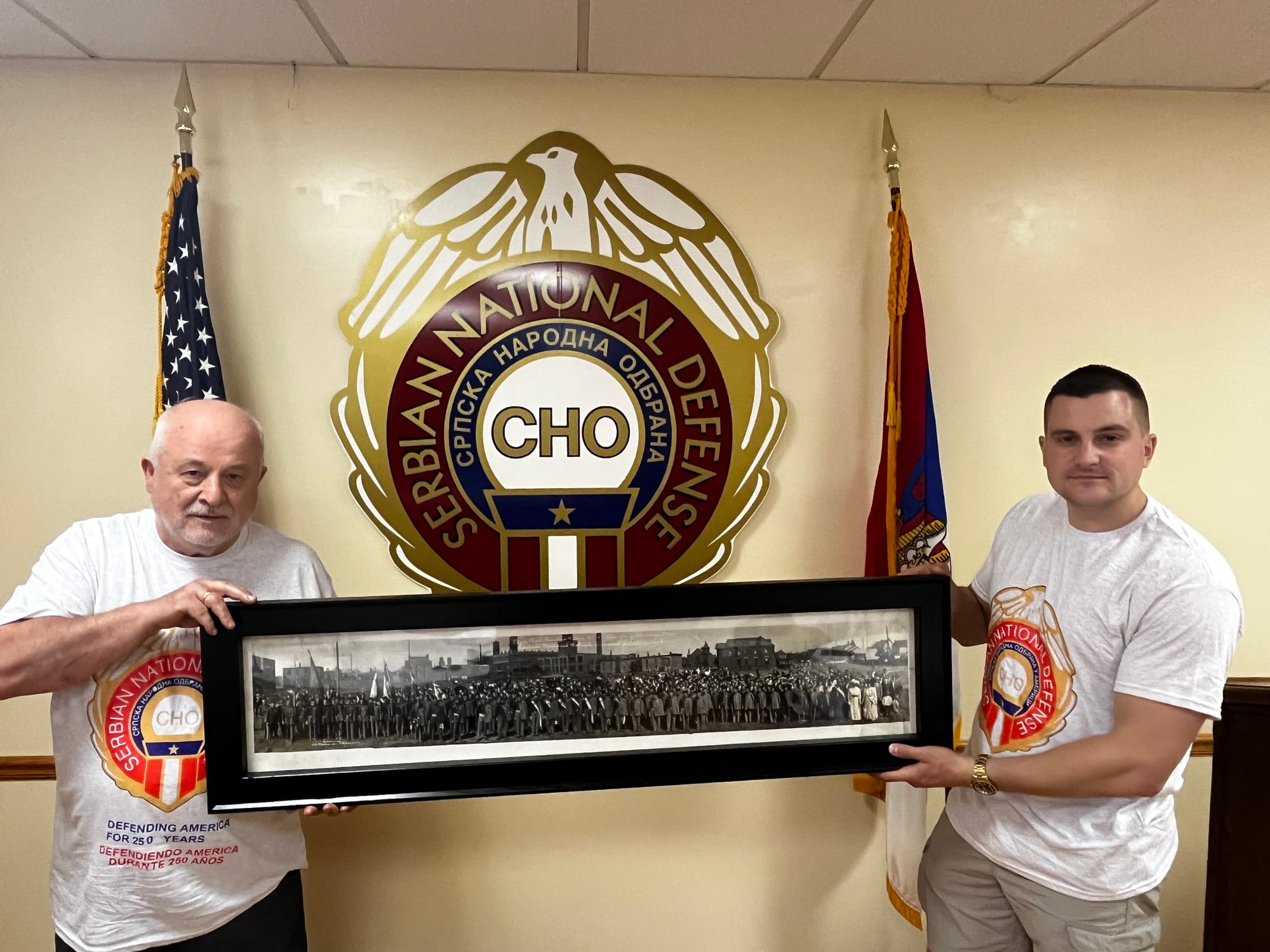 Славиша Степановић и Милан Станојевић држе историјску панорамску фотографију испред амблема Српске народне одбране у Чикагу