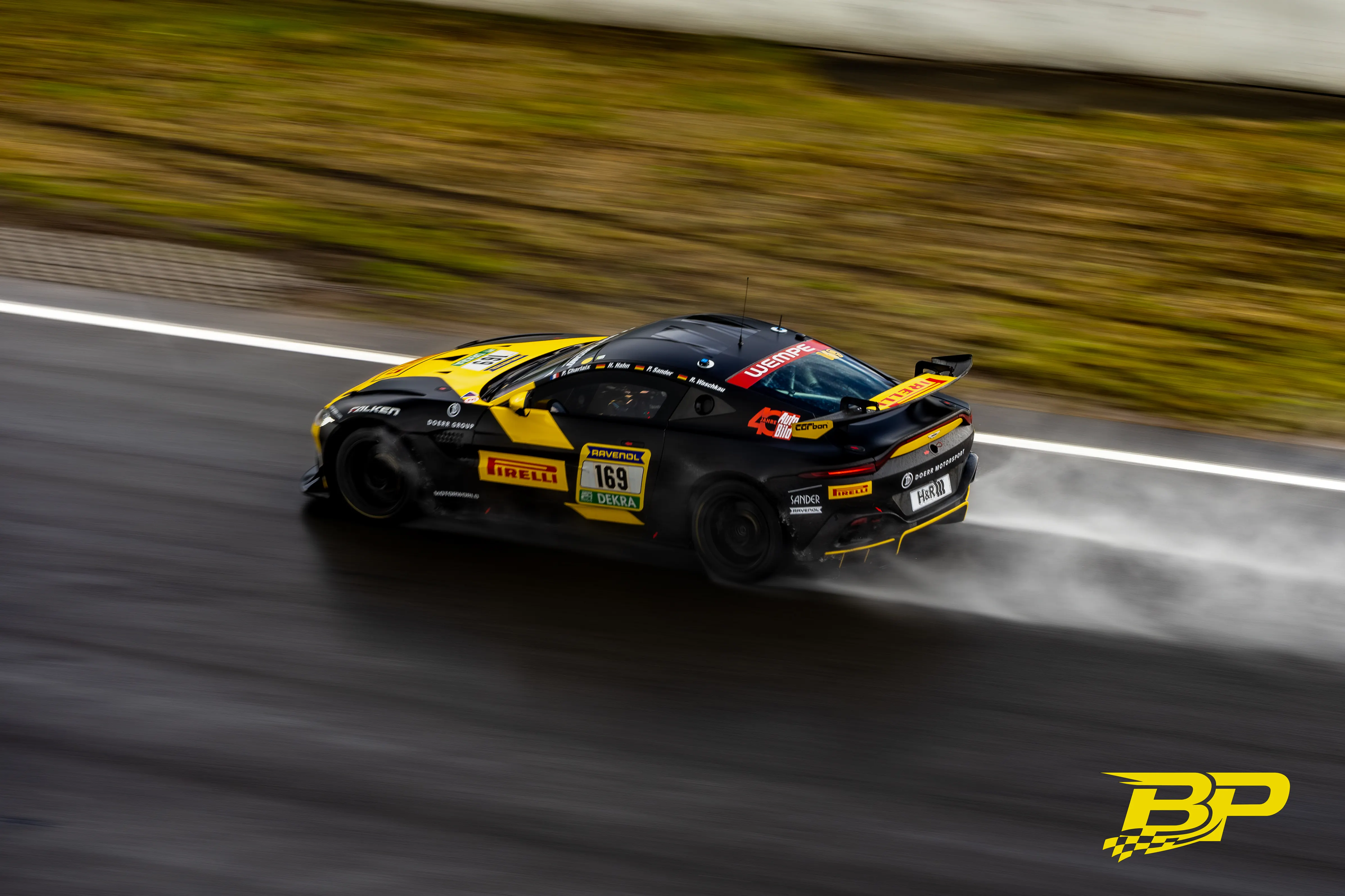 Schwarz-gelber Rennwagen mit Startnummer 169 zieht auf dem nassen Nürburgring eine Gischtfahne.