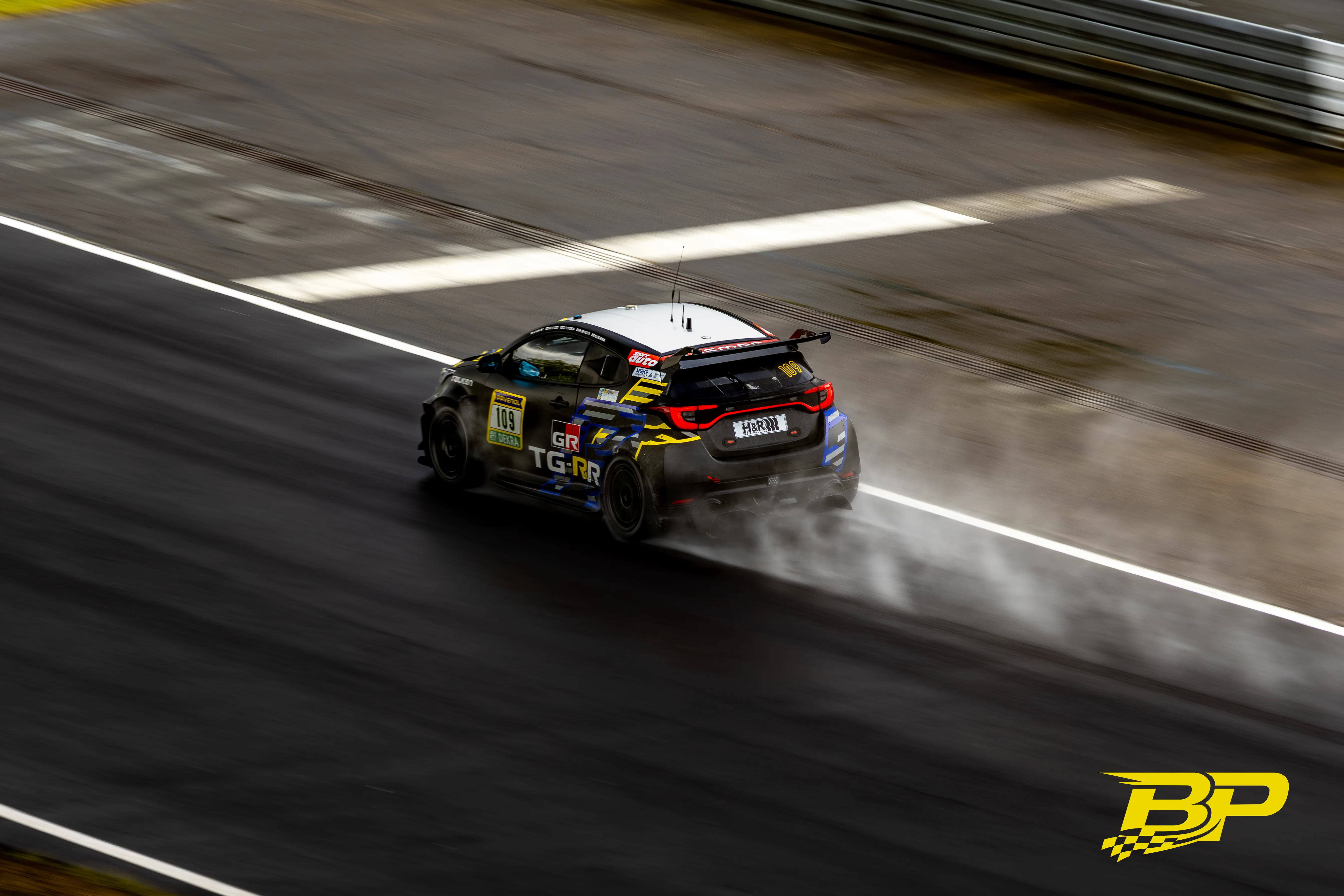 Heckansicht des schwarzen Tourenwagens mit Startnummer 109 und Gischtfahne auf nassem Asphalt.
