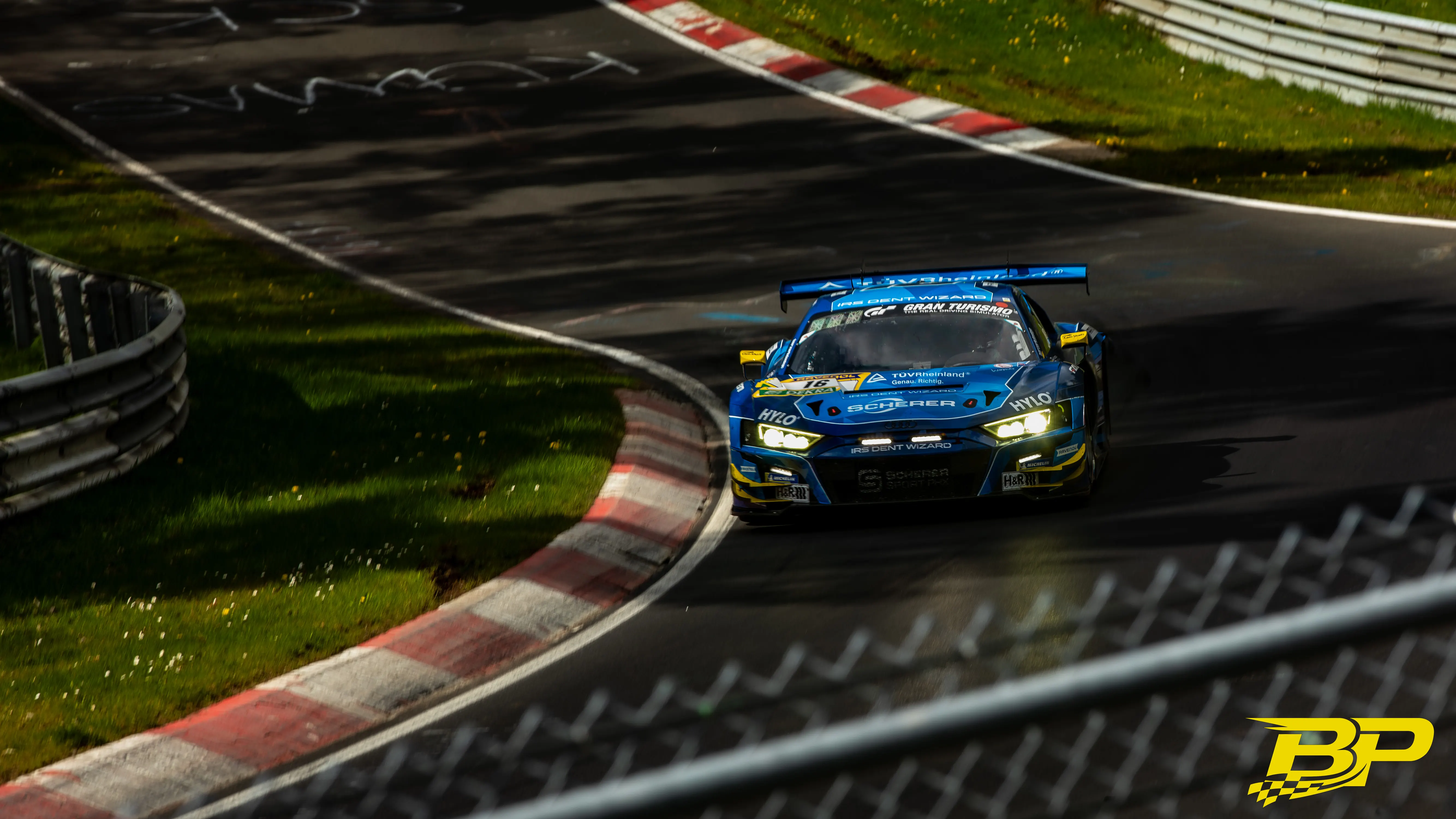 Blue GT race car with headlights on entering a shadowed forest bend.