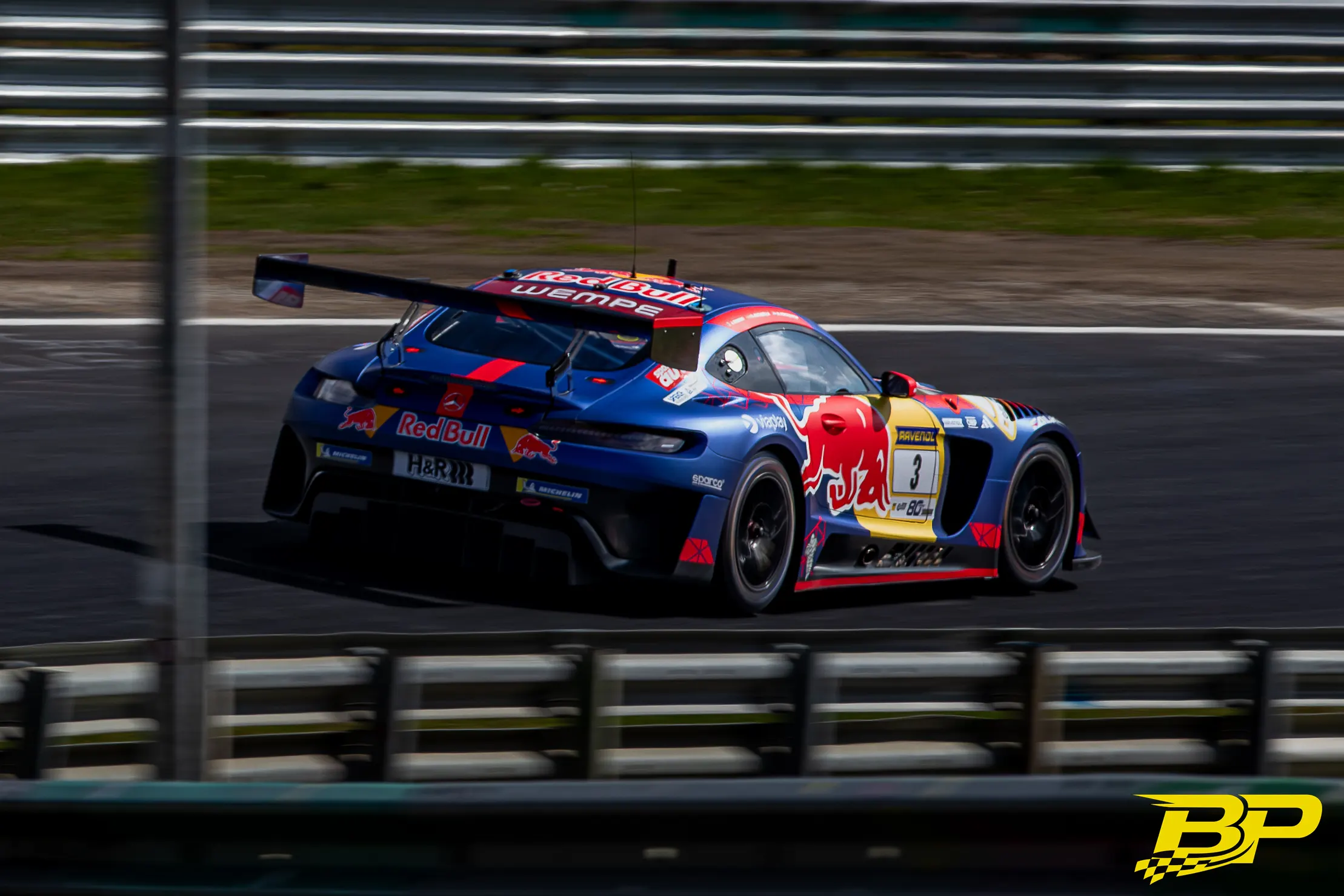 Blue-and-red Red Bull-liveried No. 3 race car speeding past Nürburgring barriers.