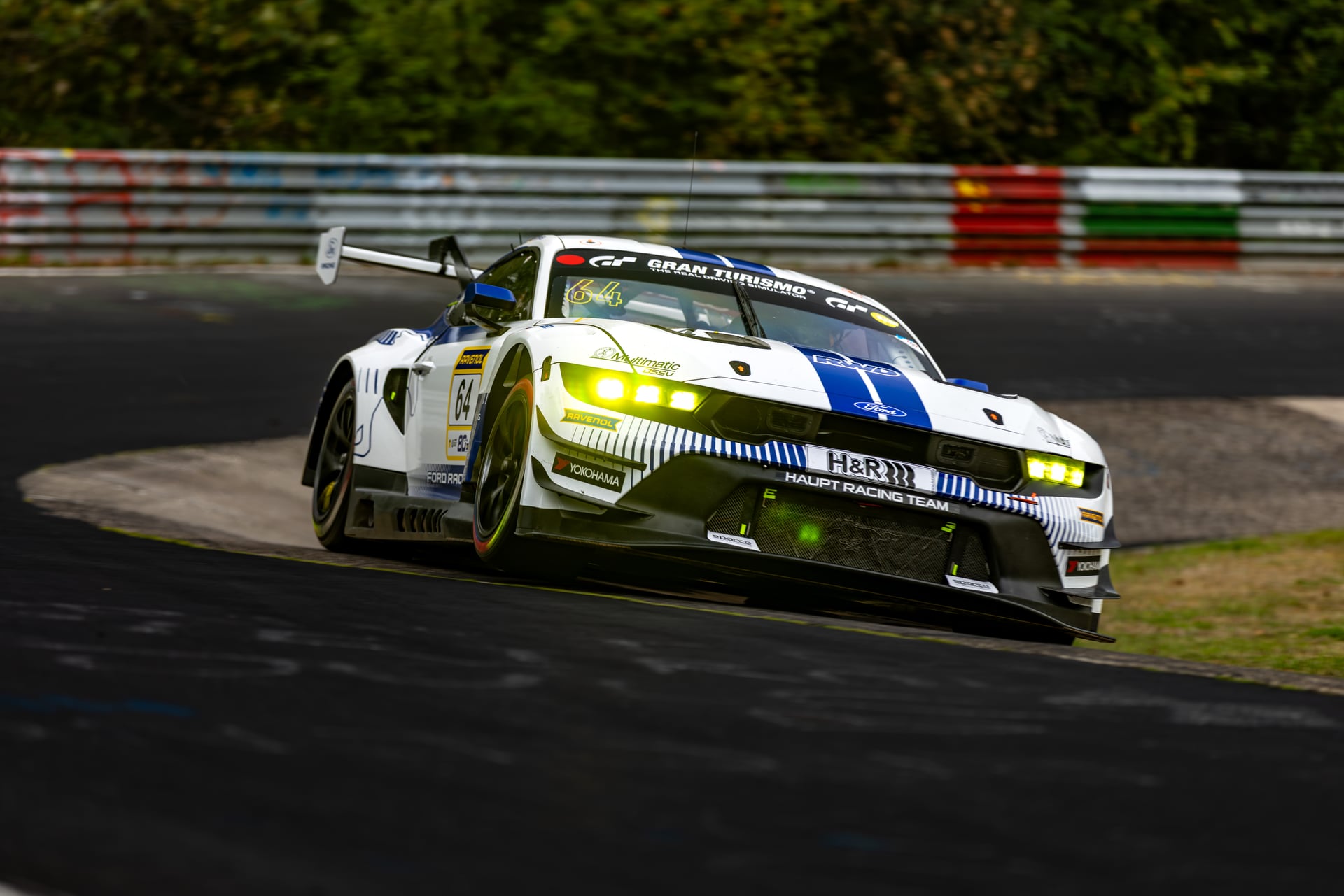 Bright green GT race car driving through a technical section of the Nürburgring with barriers and elevation changes around the track.