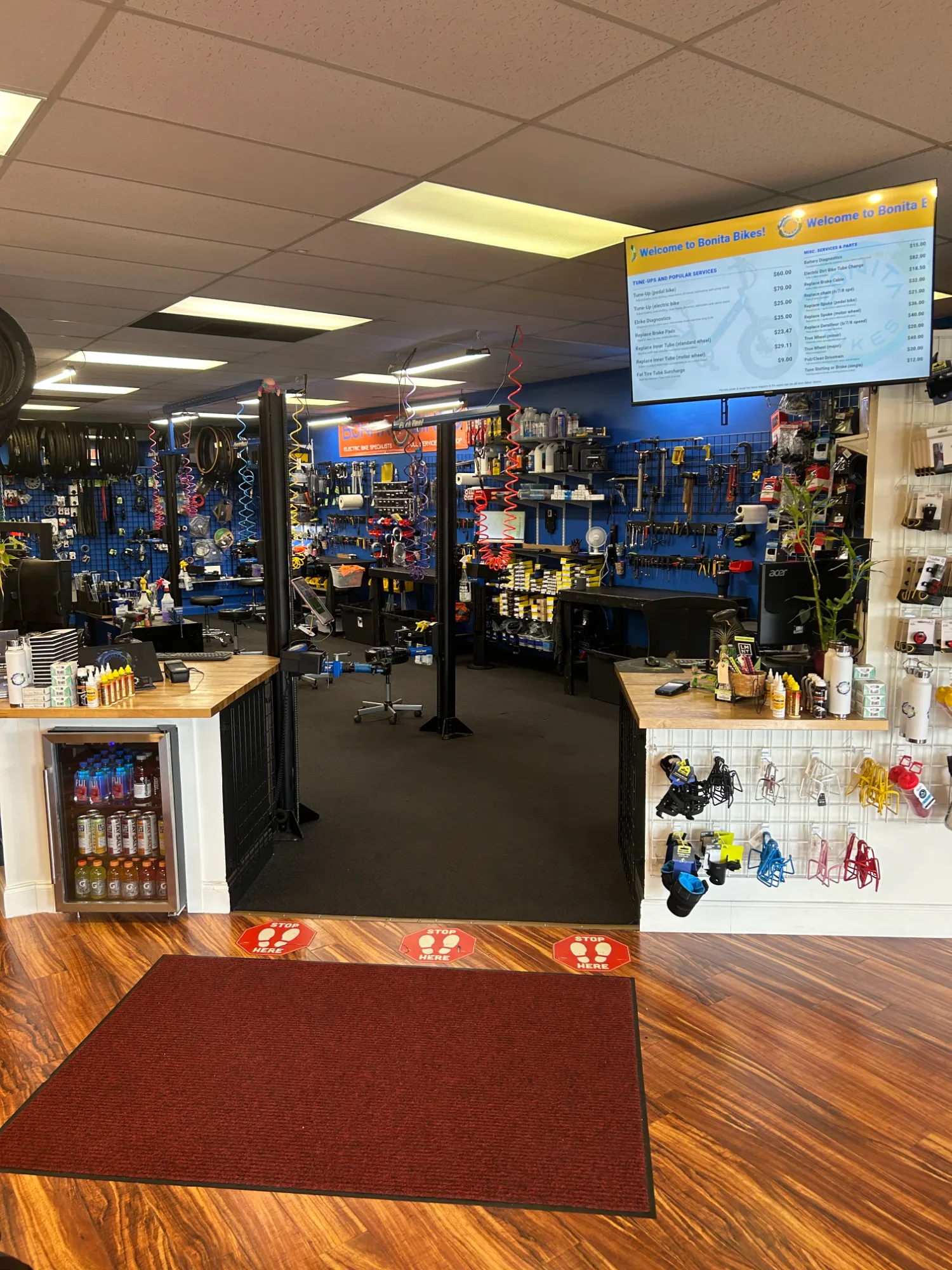 Inside Bonita Bikes shop entrance looking toward the service counter, with a digital price list display overhead, beverage cooler, and water bottle cages and bike accessories on grid wall displays