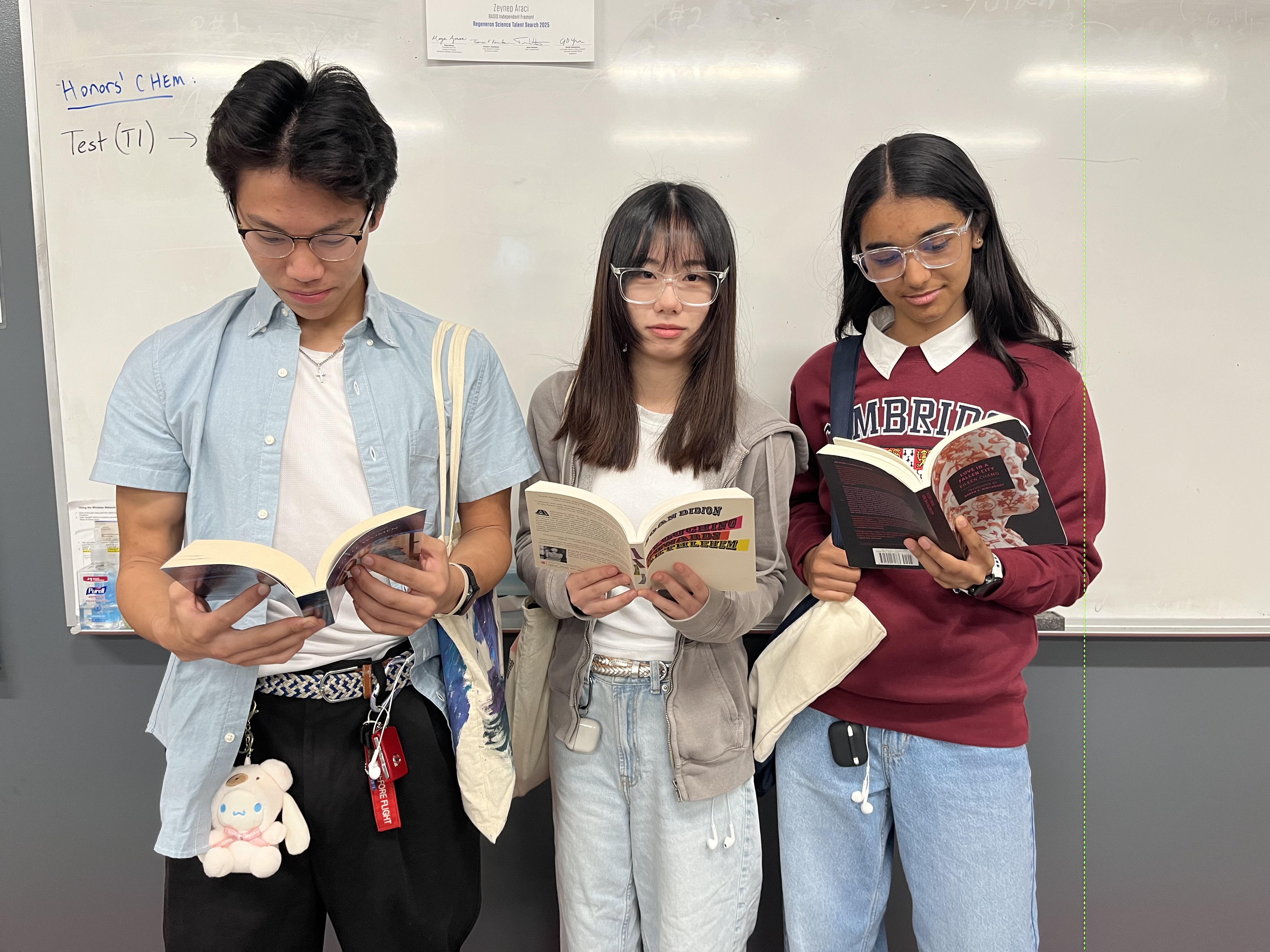 Three students stand in front of a whiteboard while holding books.