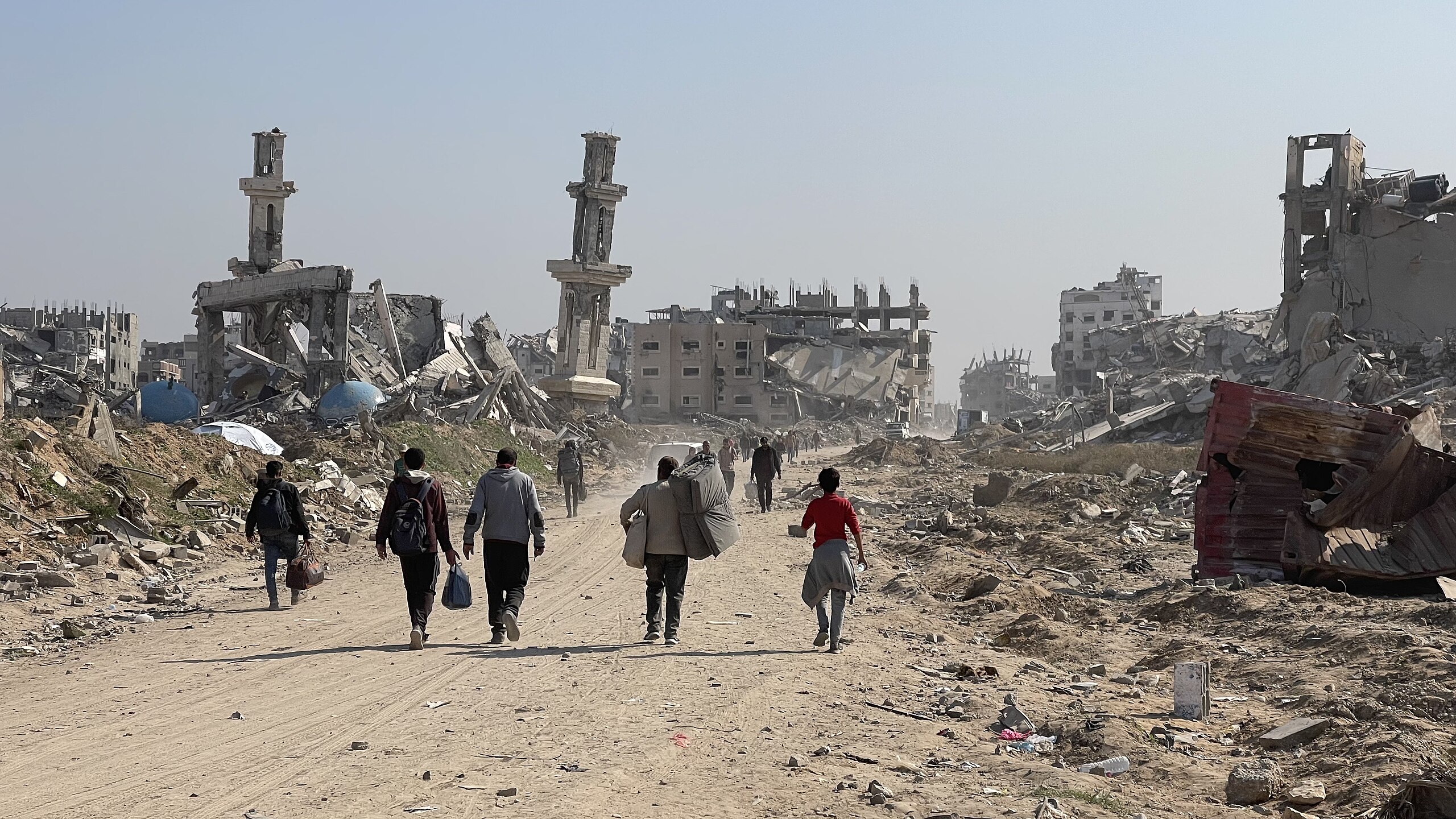 People walk along a dirt road between heavily damaged and collapsed buildings and rubble.