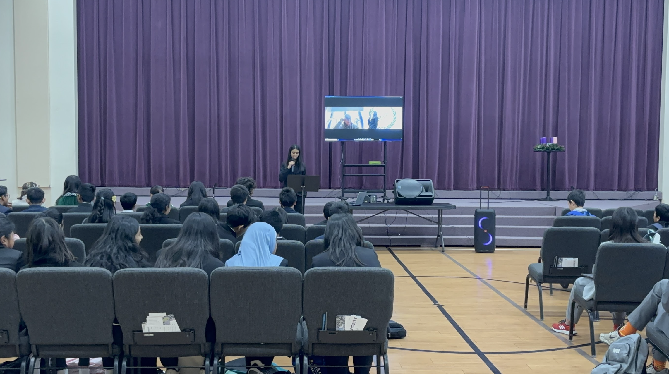 Person speaking on a stage in front of an audience seated in a school multipurpose room.
