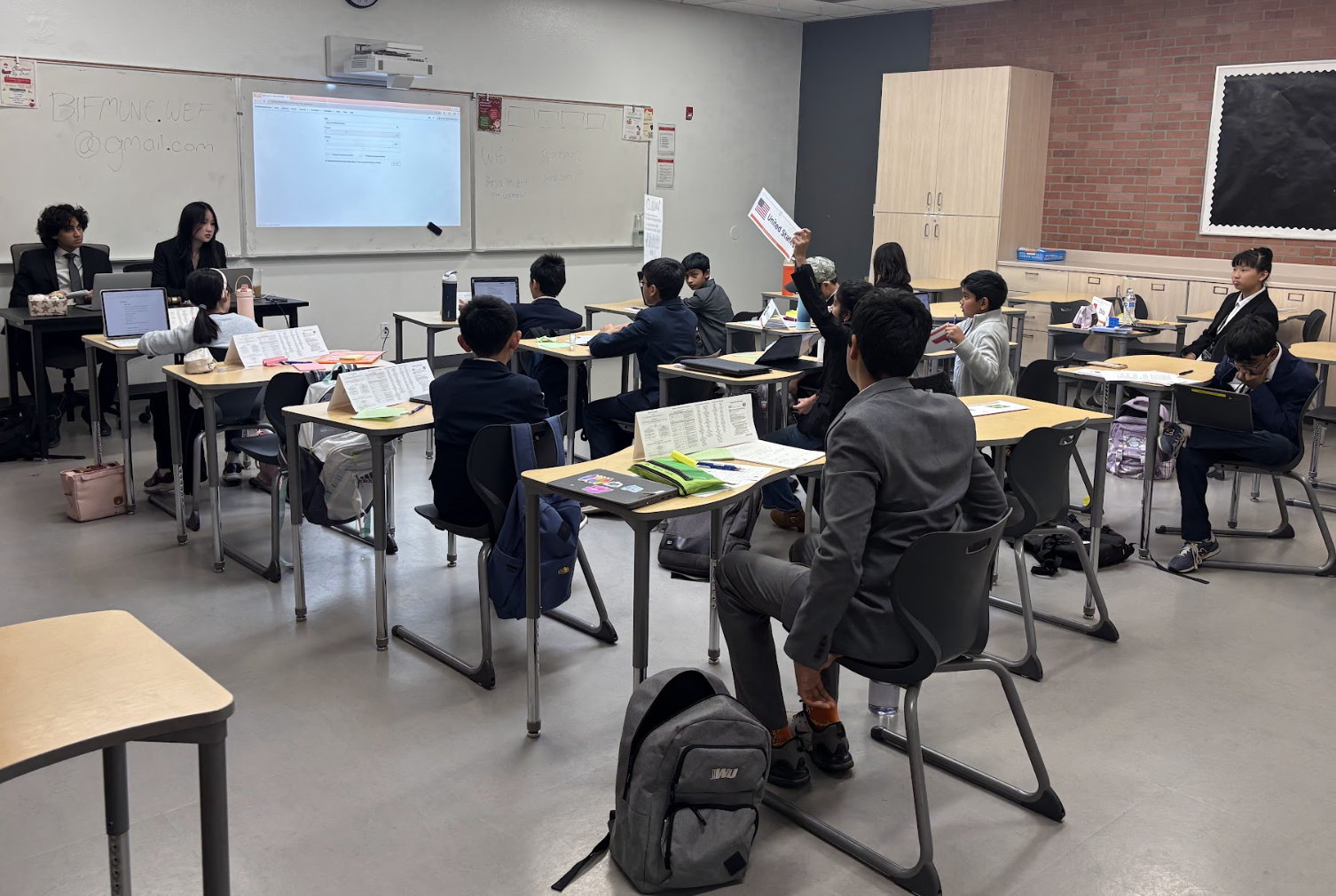 Students seated at desks in a classroom while several people sit at the front.