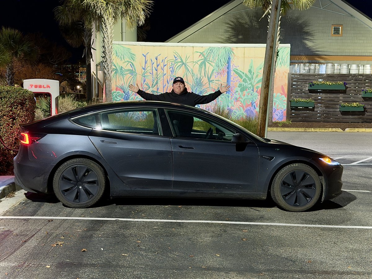 A person stands with arms outstretched beside a gray Tesla Model 3 in a parking space.