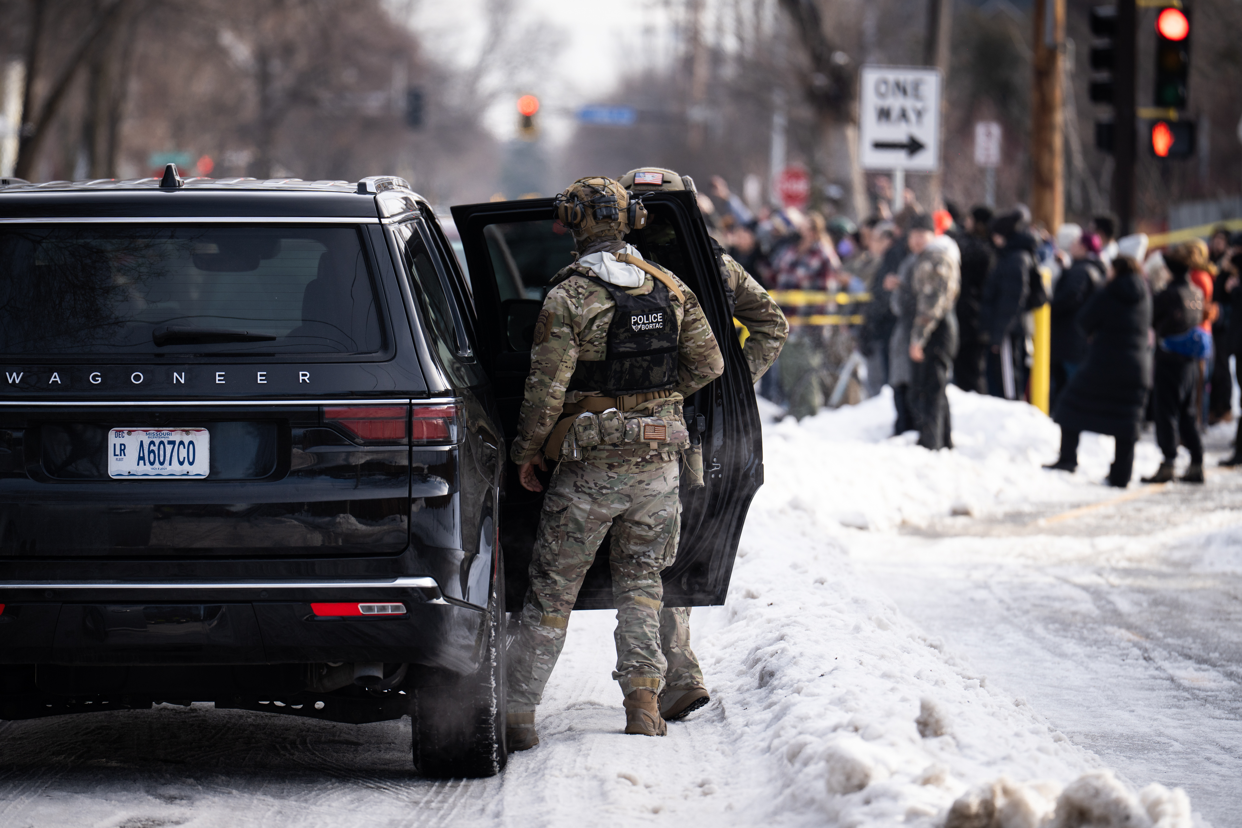 Armed officer standing beside an open vehicle door on a snowy city street, with people in the background.
