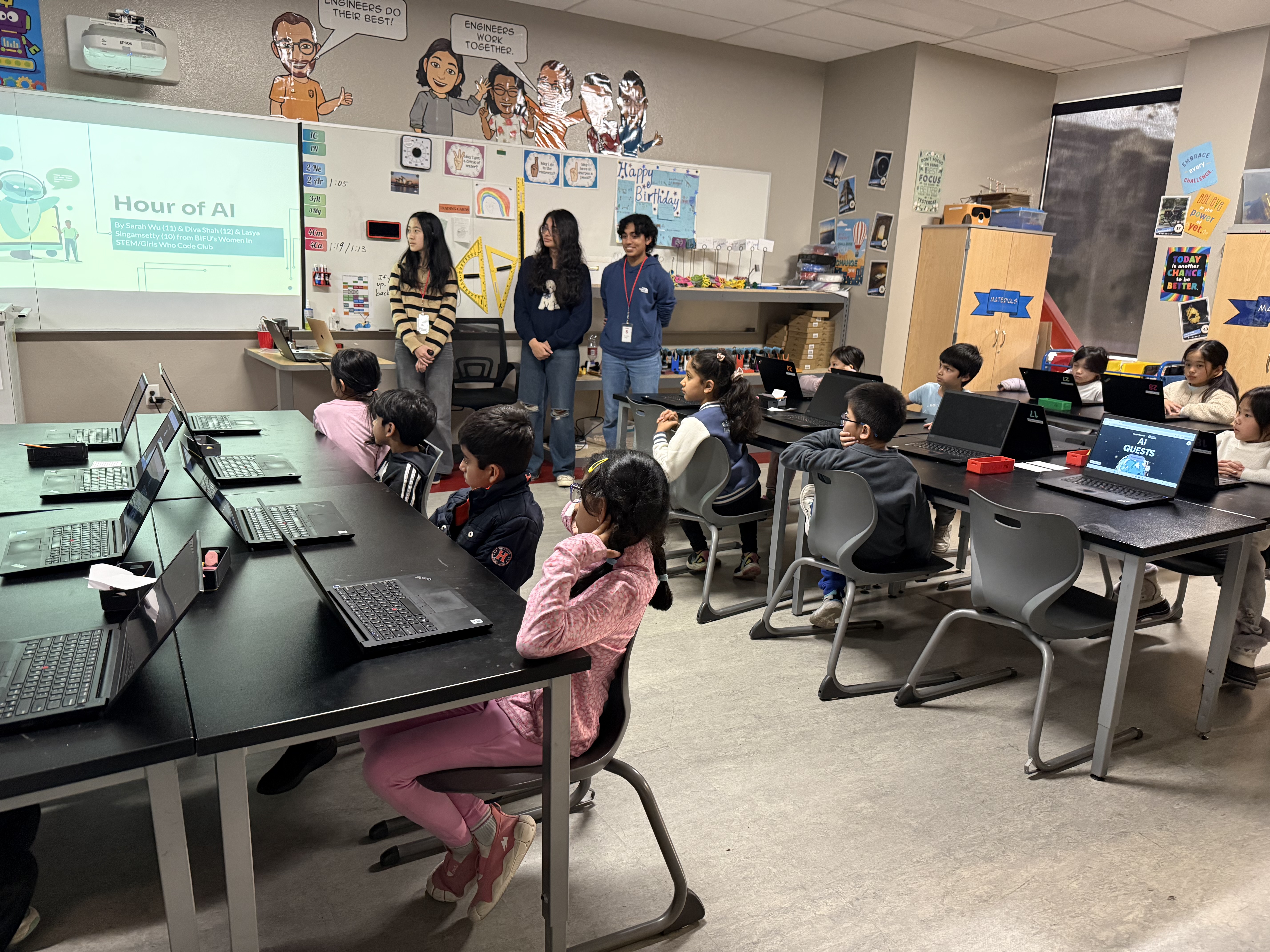 Students seated at computers in a classroom while instructors stand near an Hour of AI presentation.