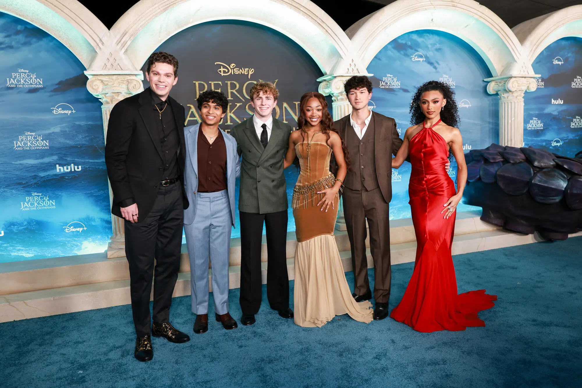 Six people in formal clothing pose on a blue carpet in front of Percy Jackson promotional displays.