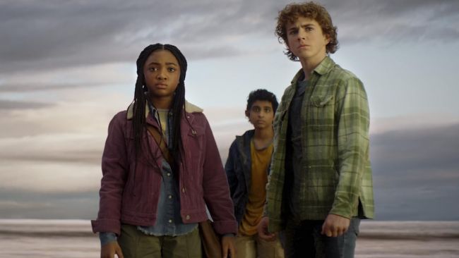 Three people stand outdoors near a beach under a cloudy sky.