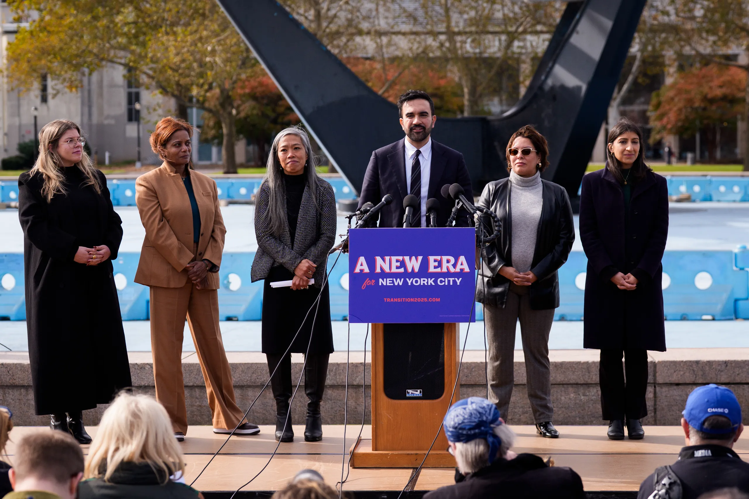 Zohran Mamdani stands at a podium with several women during an outdoor event.