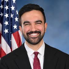 Portrait of Zohran Mamdani in a suit with an American flag behind him.