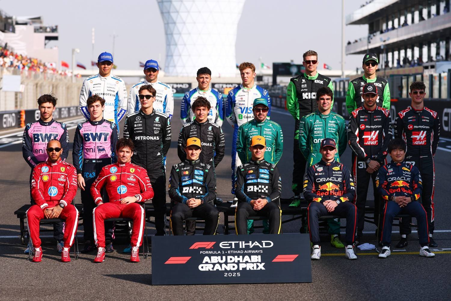 Group of Formula 1 drivers seated and standing together beside a 2025 Abu Dhabi Grand Prix sign.