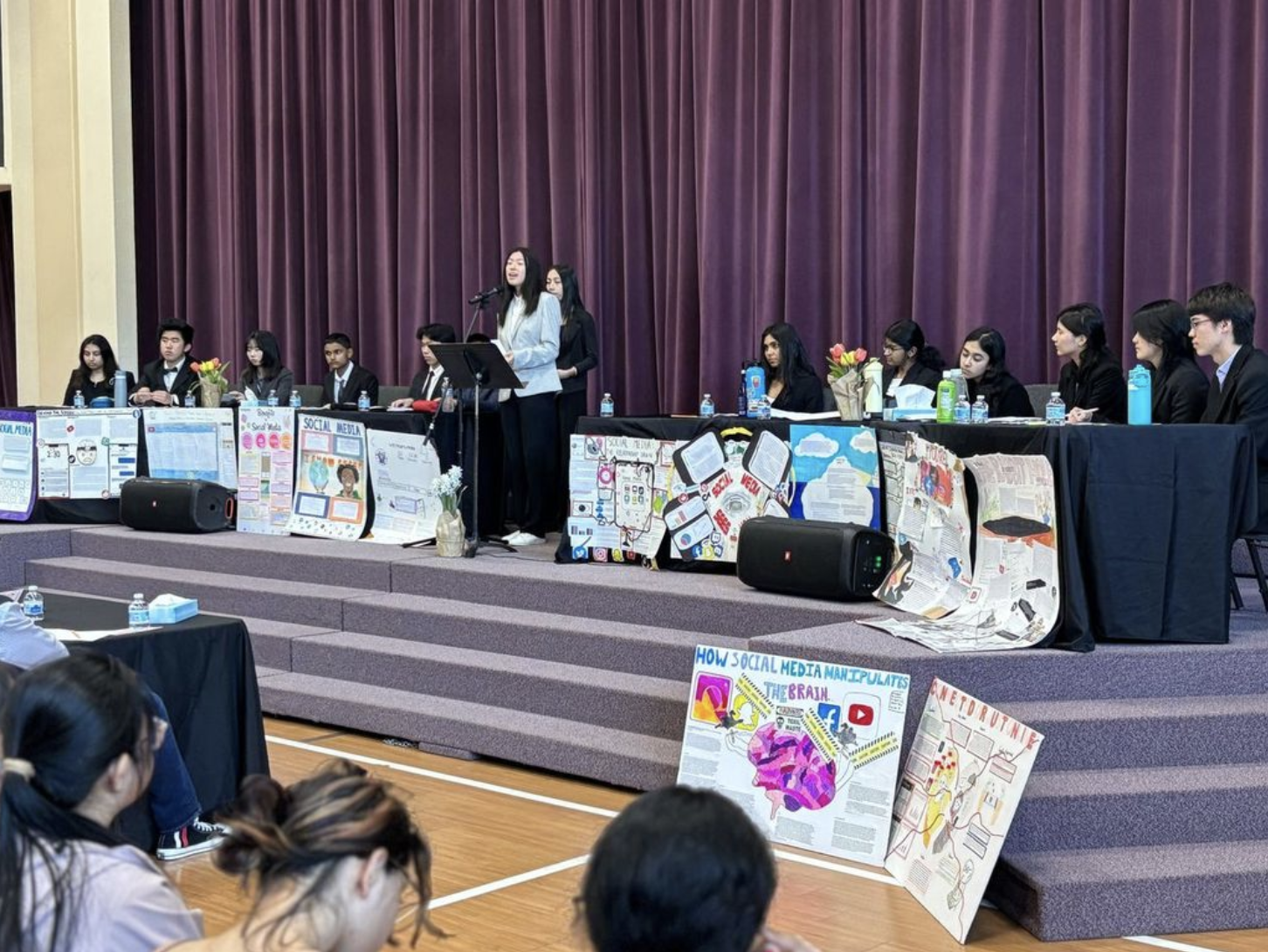 Students speak at a debate podium while teams sit behind tables displaying social media posters.