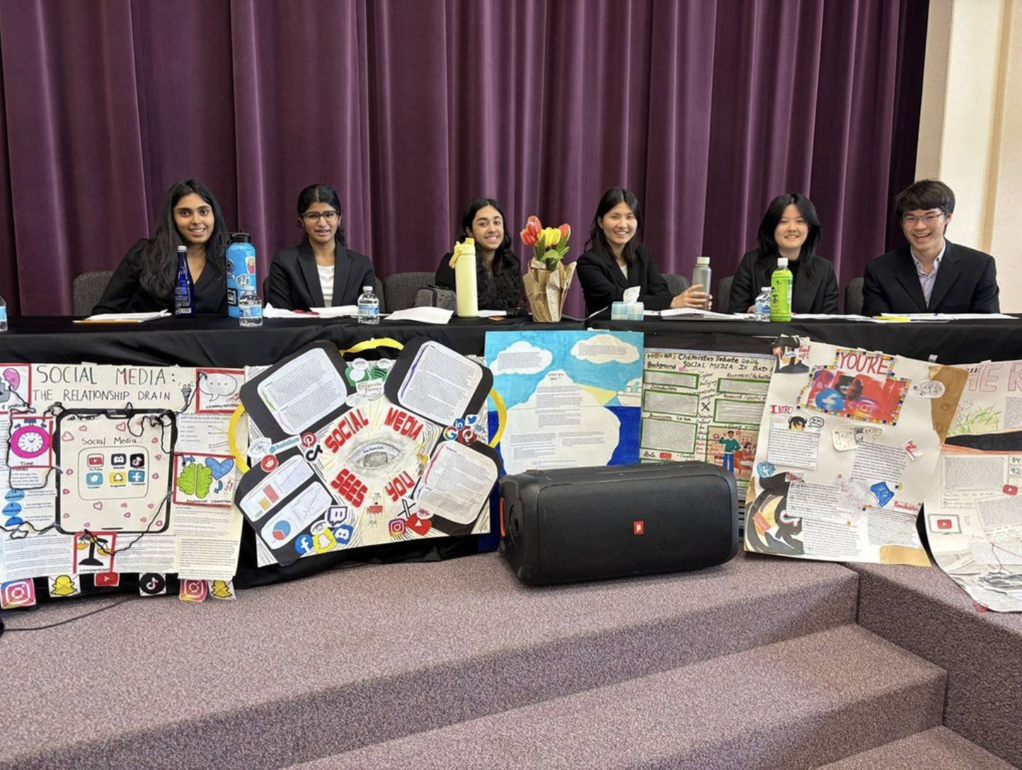 Six students seated behind a table with social media debate posters and a speaker.