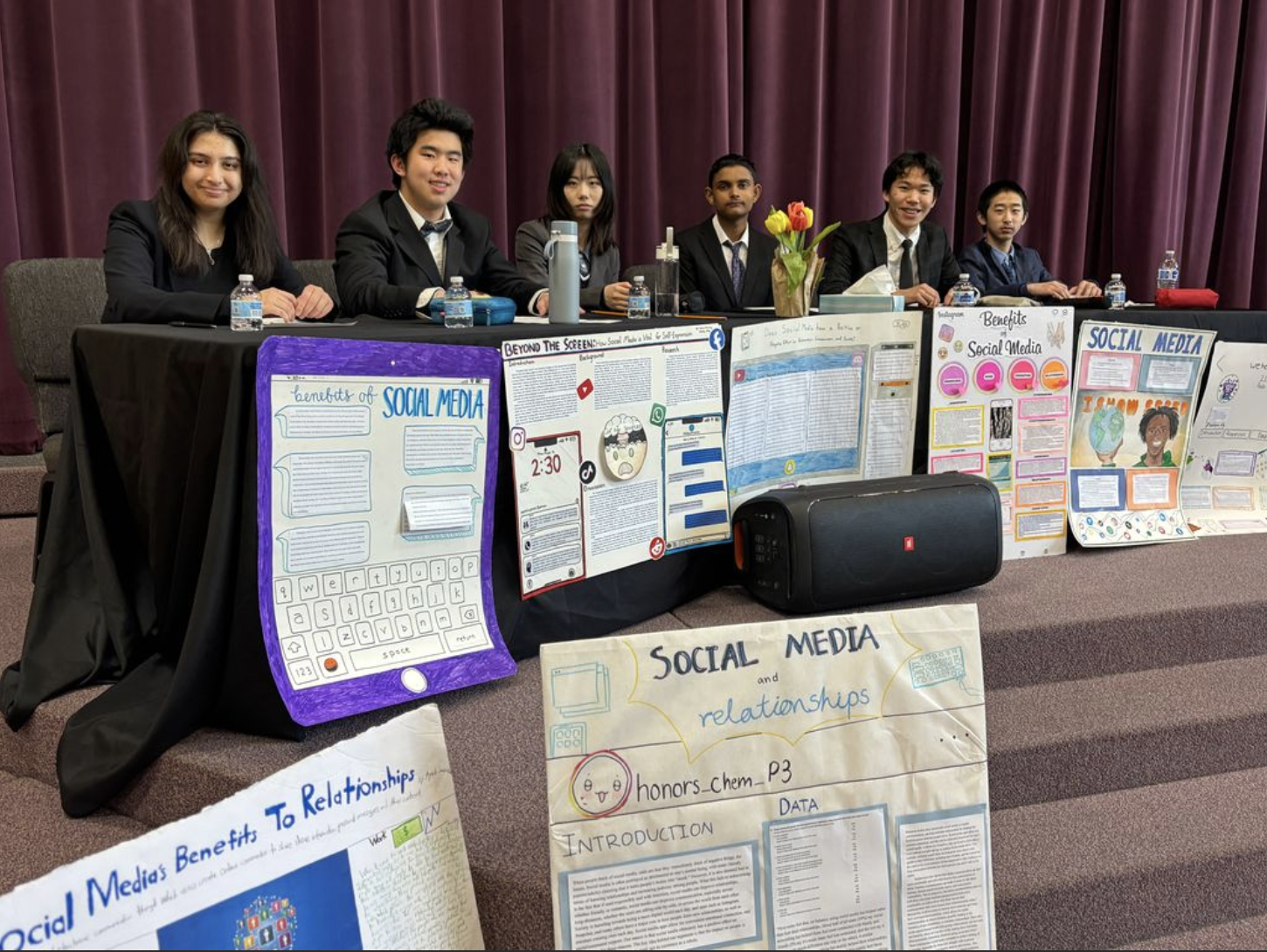 Six students seated behind a table with social media debate posters and a speaker.