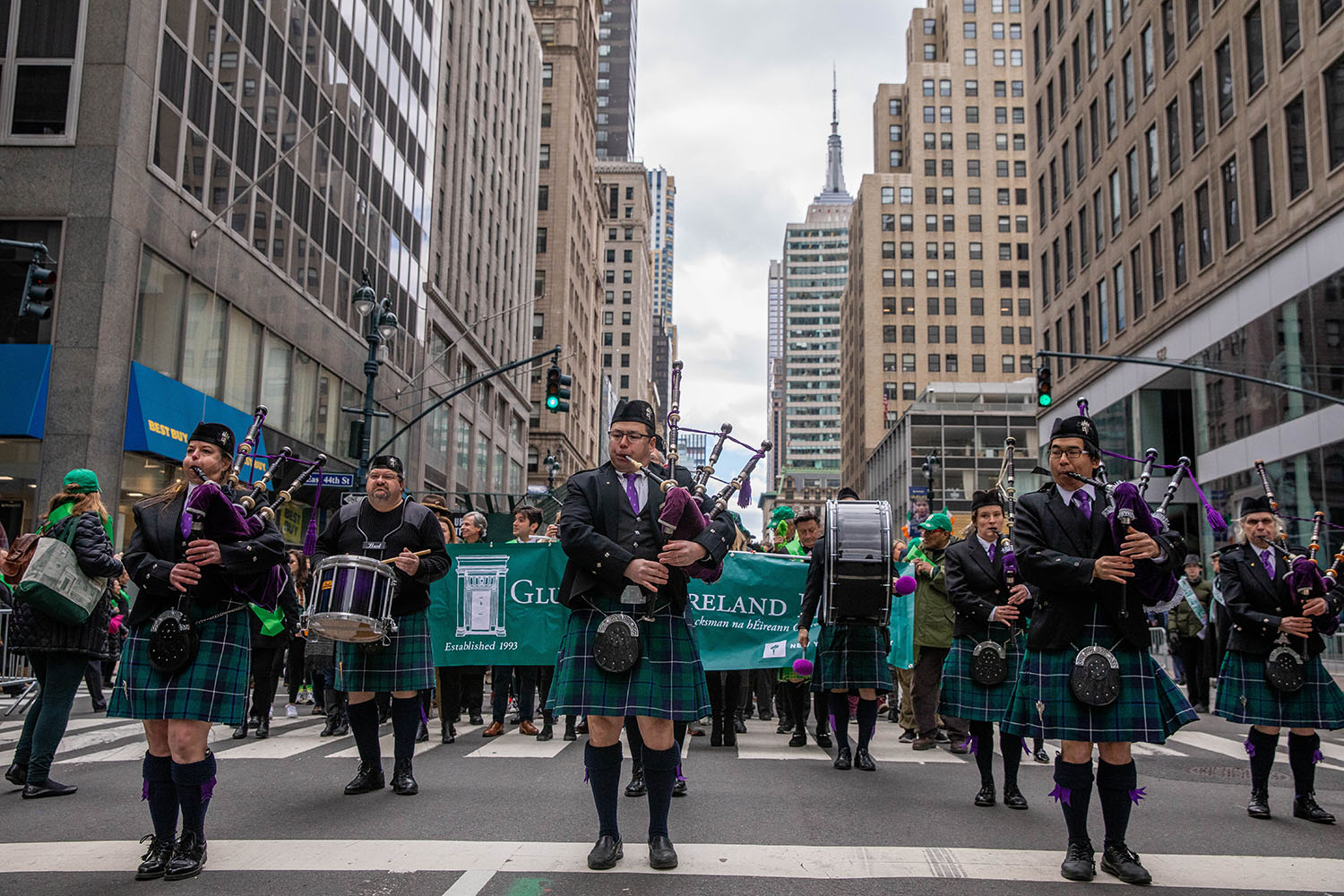 Marching musicians in kilts and purple-and-green clothing lead a St. Patrick’s Day parade down a city street.