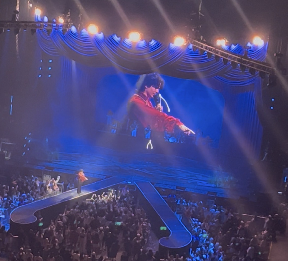 Performer on a blue-lit stage above a crowd, with a wishbone-shaped catwalk extending into the audience.