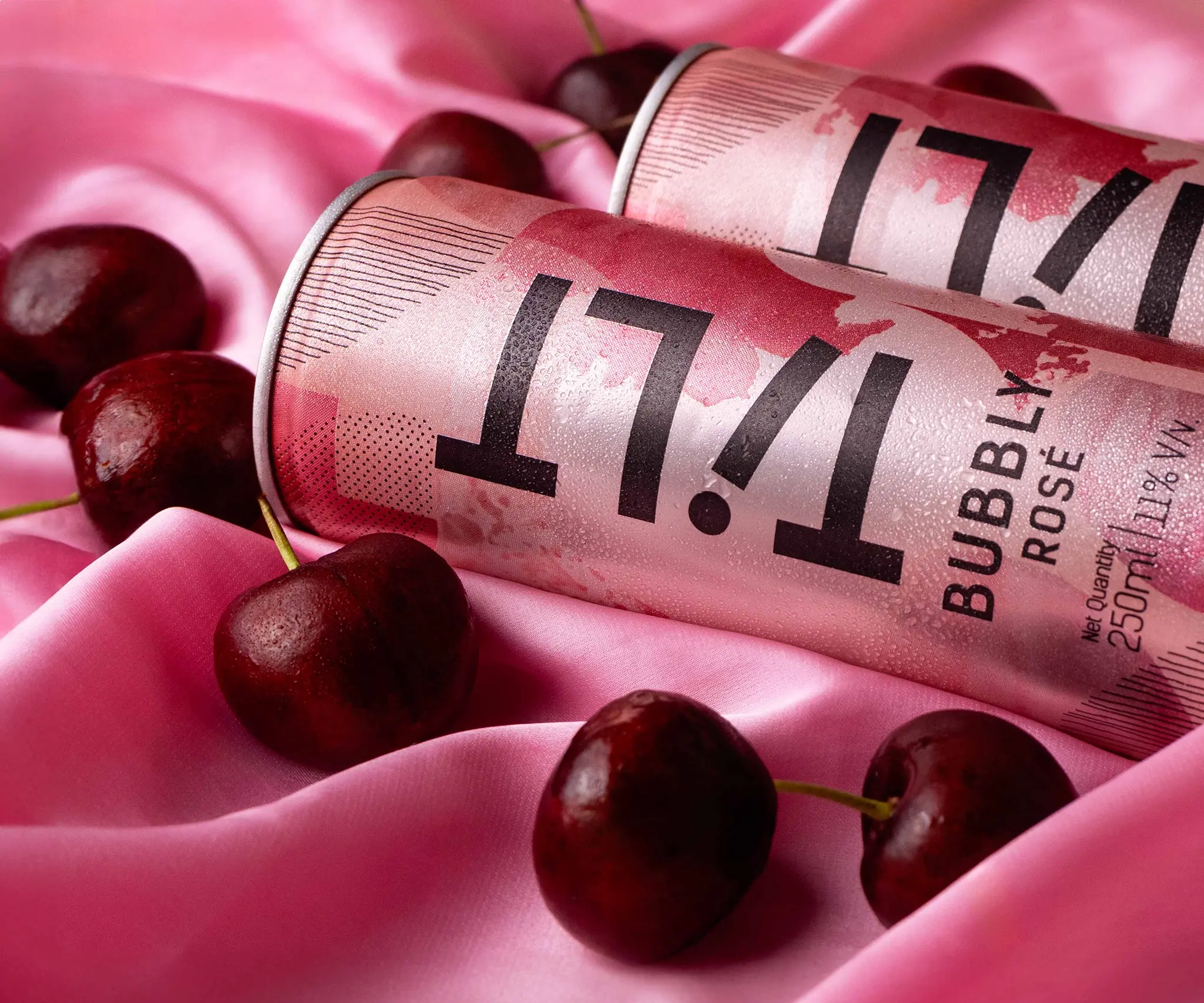 Top angle shot of two wine cans by tilt, romantic set styling with pink silk fabric and cherries scattered on set, soft diffused highlights and shadows