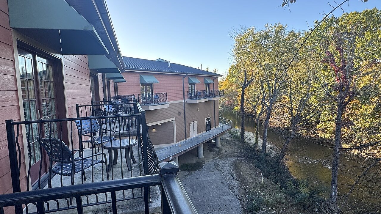A hotel balcony with black metal railings and chairs overlooking a river lined with autumn trees and a multi-storey hotel building.