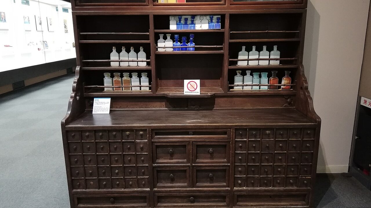 A dark wooden apothecary cabinet with small drawers and shelves holding white and blue glass medicine bottles, in a museum display.