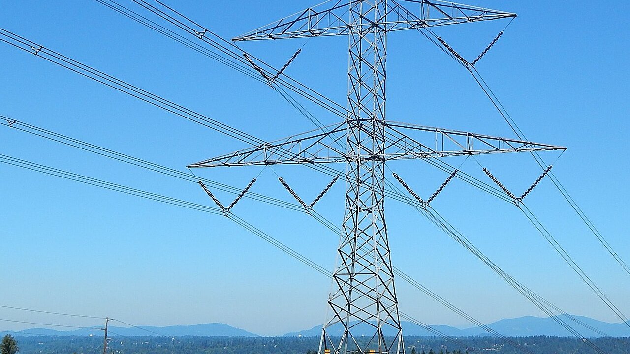 A tall steel high-voltage transmission tower carrying power lines against a blue sky, with hills in the distance.
