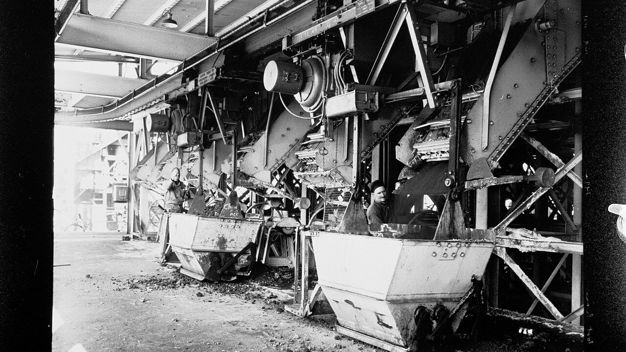 Two workers beside large industrial machinery with metal hoppers and conveyors in a black and white photograph of a plant