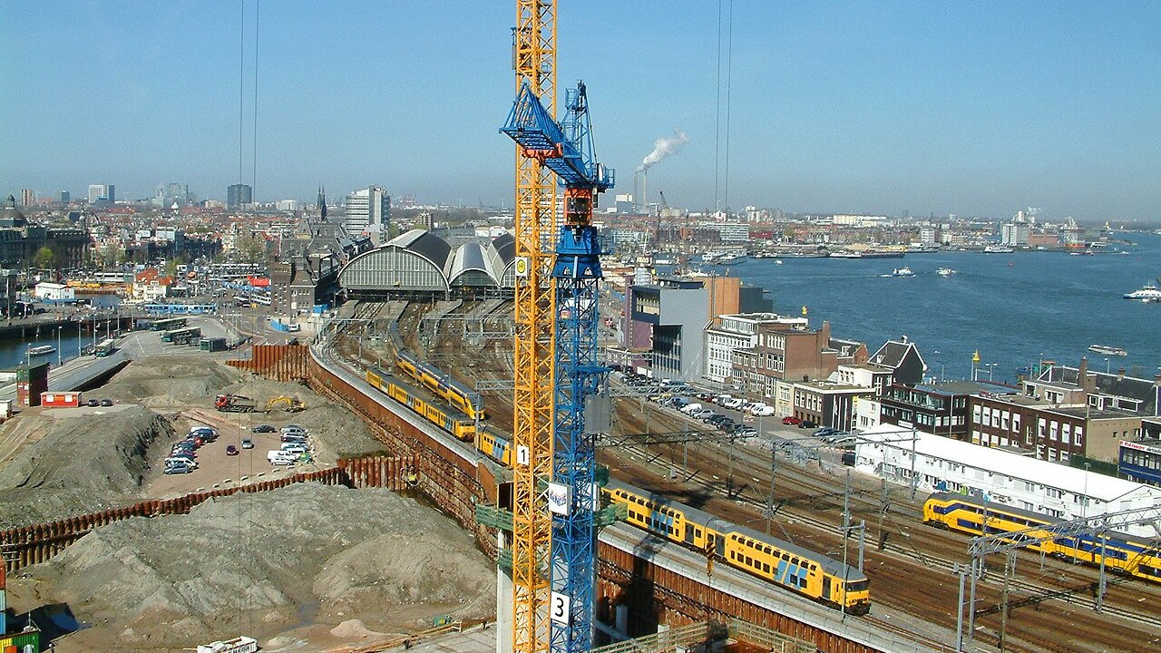 A construction site with tower cranes and excavation beside a railway station, with yellow trains on the tracks and a harbour behind.