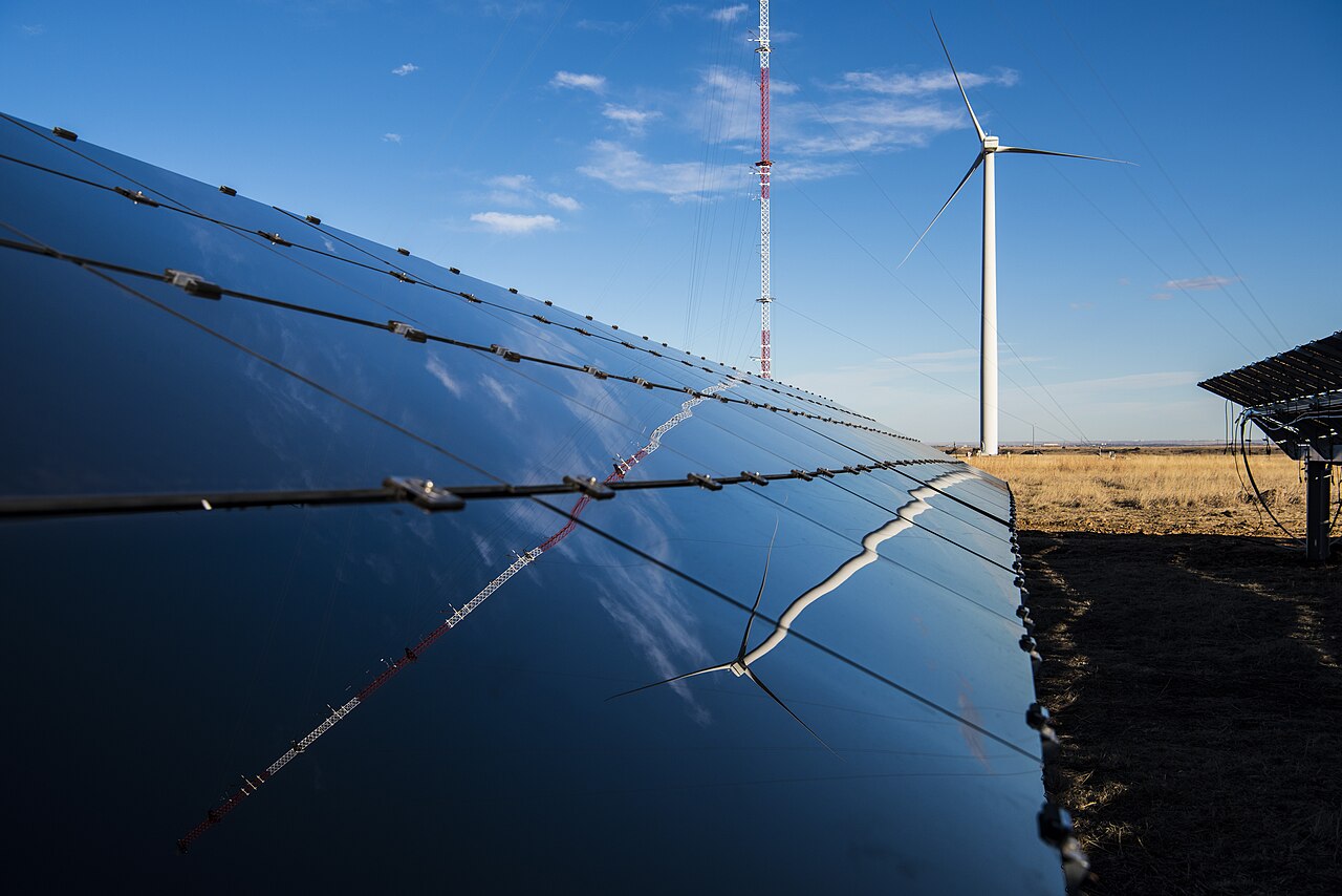Solar panels in the foreground with a wind turbine and a mast behind them