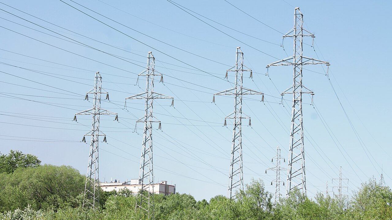 Several tall steel lattice electricity transmission towers carrying power lines across a green landscape under a clear sky.