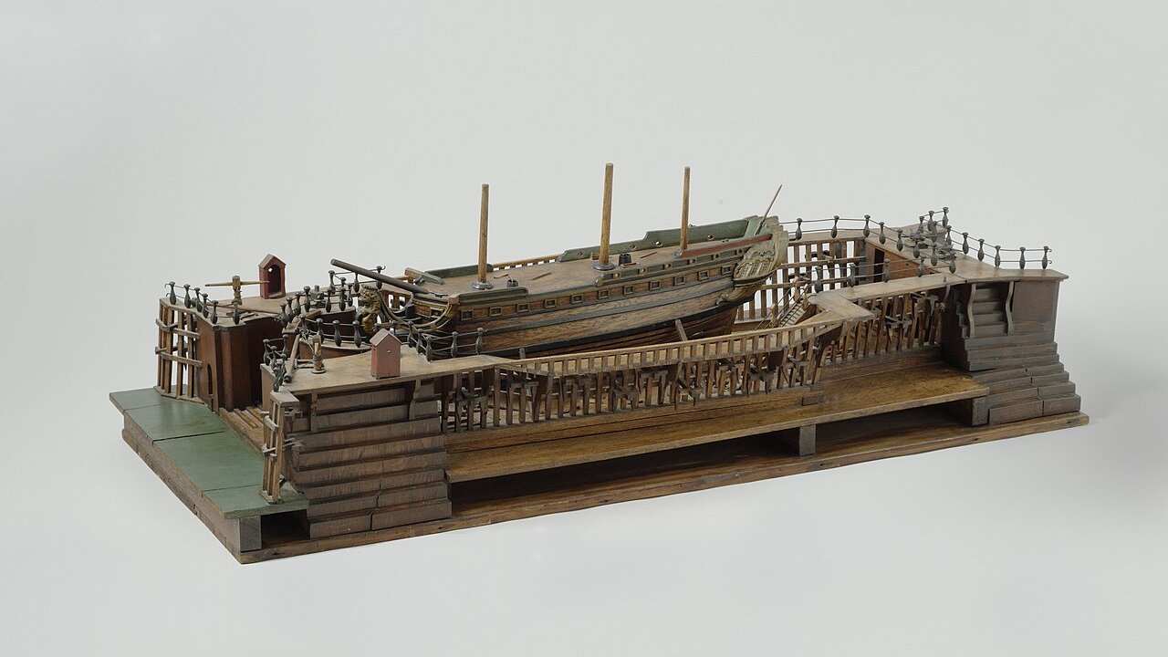 A wooden model of a dry dock with a sailing ship in it, shown on a plain background.