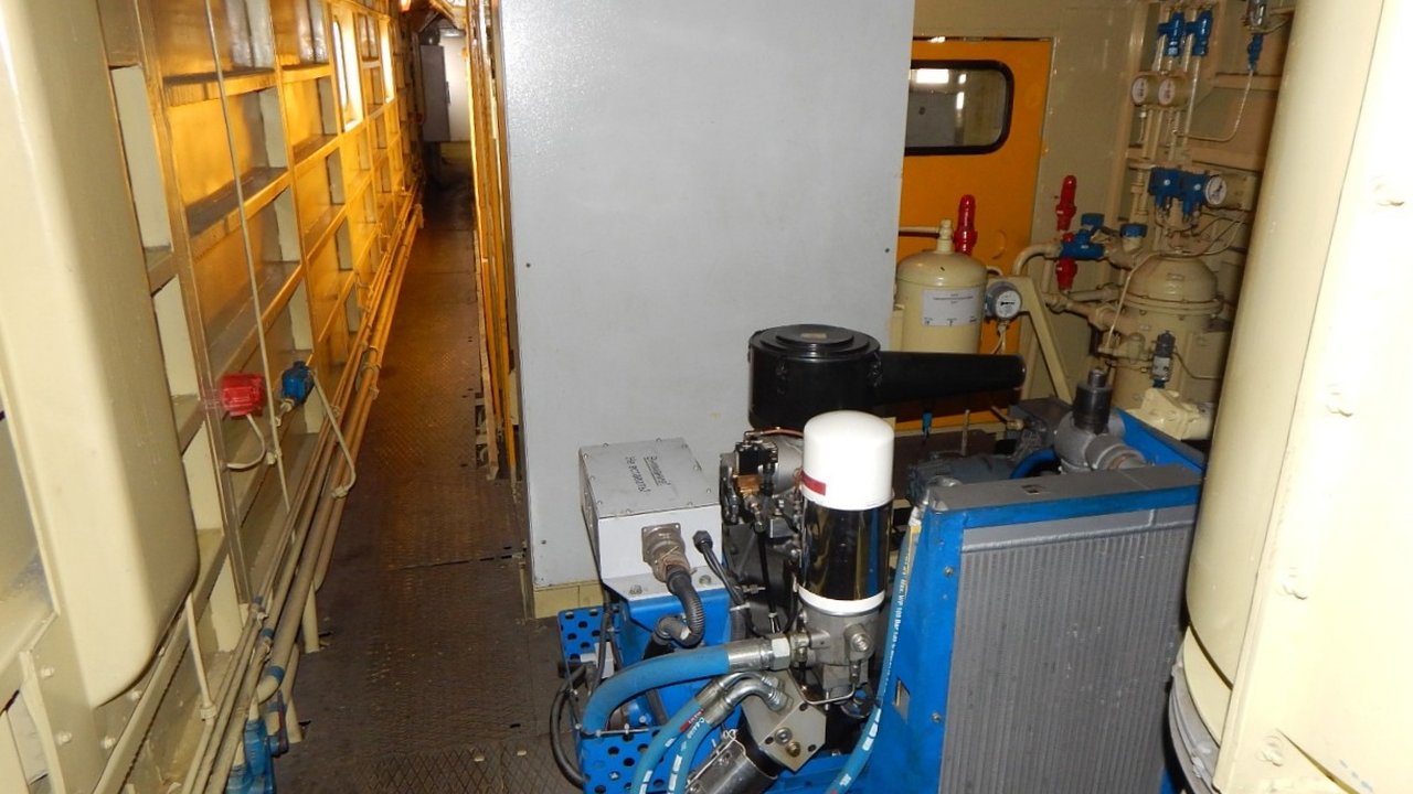 An air compressor unit on a blue skid beside pipes and gauges in a narrow machinery room.