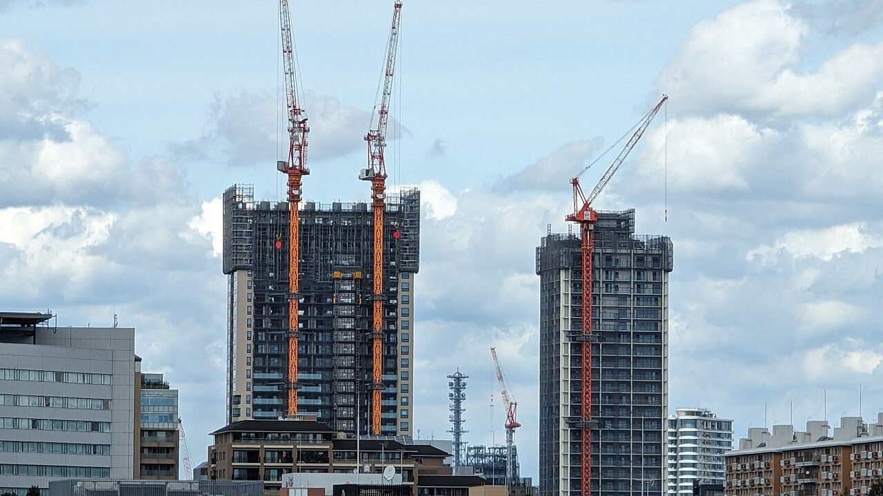 Two high-rise residential towers under construction, wrapped in scaffolding and served by red and orange tower cranes against a cloudy sky.