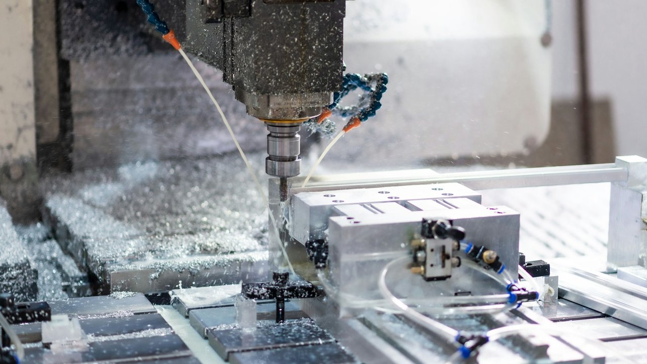 A CNC machine spindle milling an aluminium block, with coolant lines spraying and metal chips on the bed.