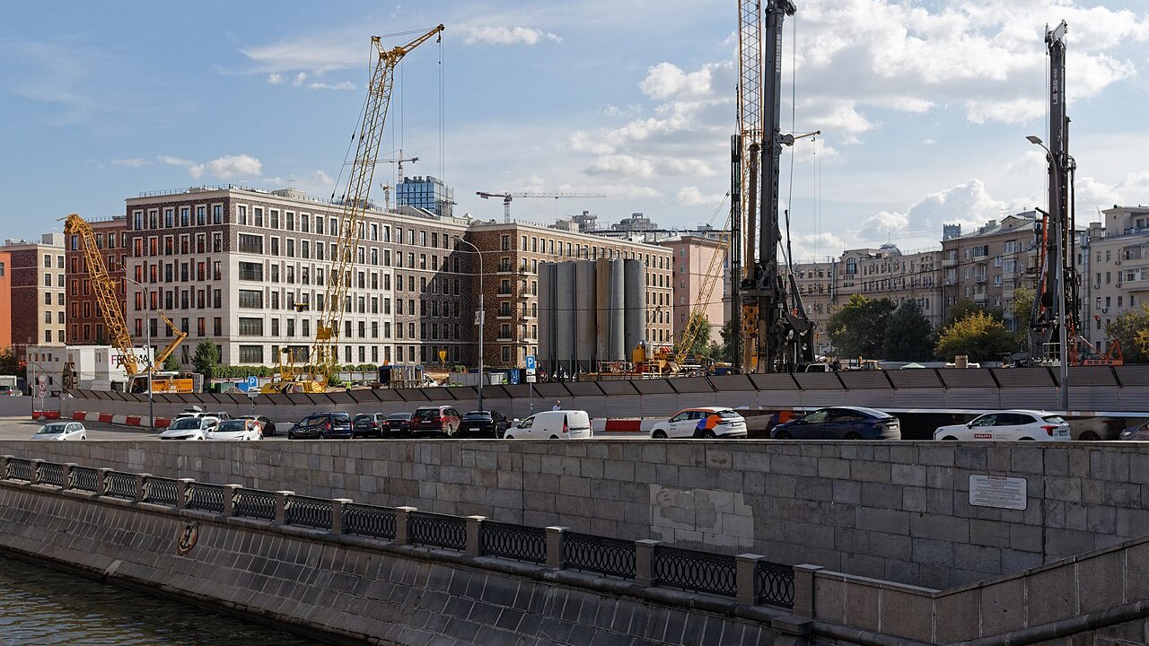 Cranes and piling rigs working on a city construction site beside a riverside road with cars and new multi-storey buildings behind.