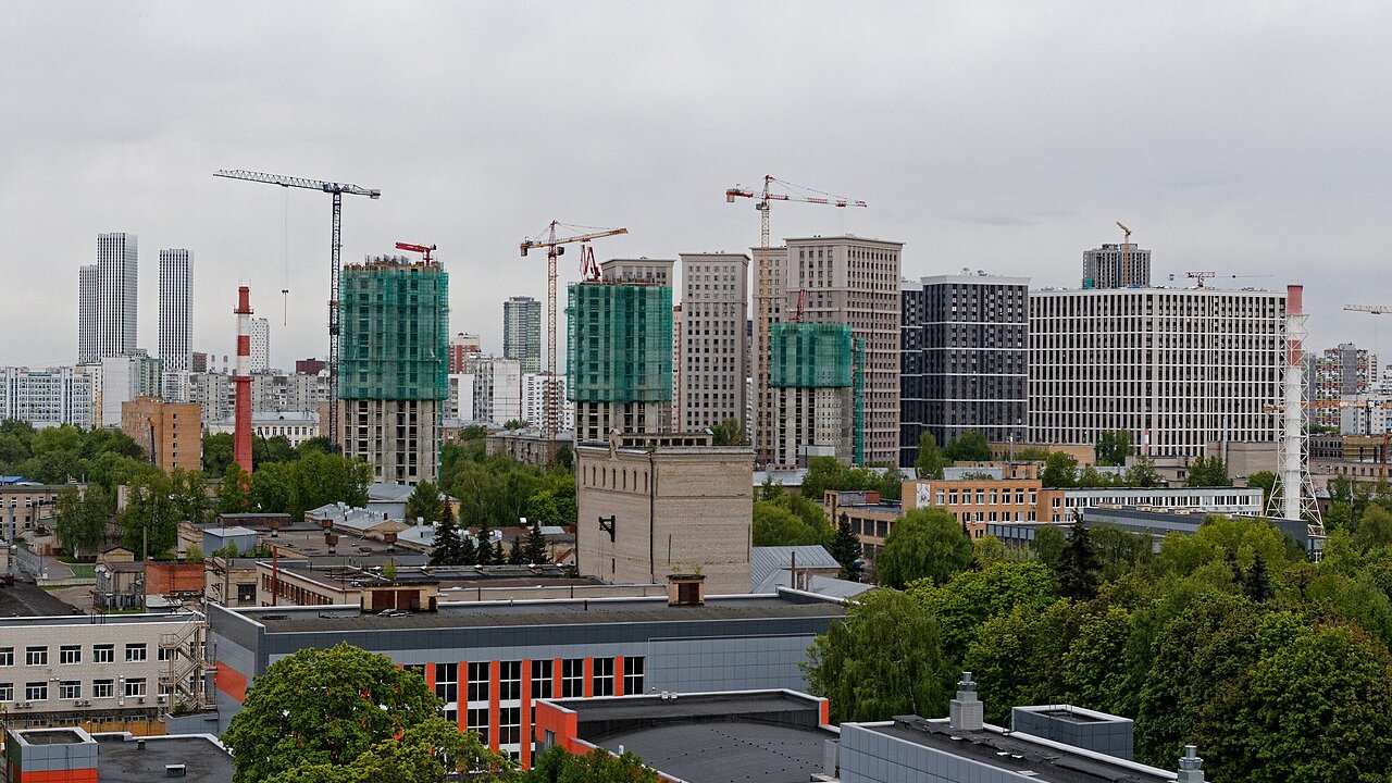 A city skyline of high-rise towers, several under construction with cranes and green safety netting, under an overcast sky.