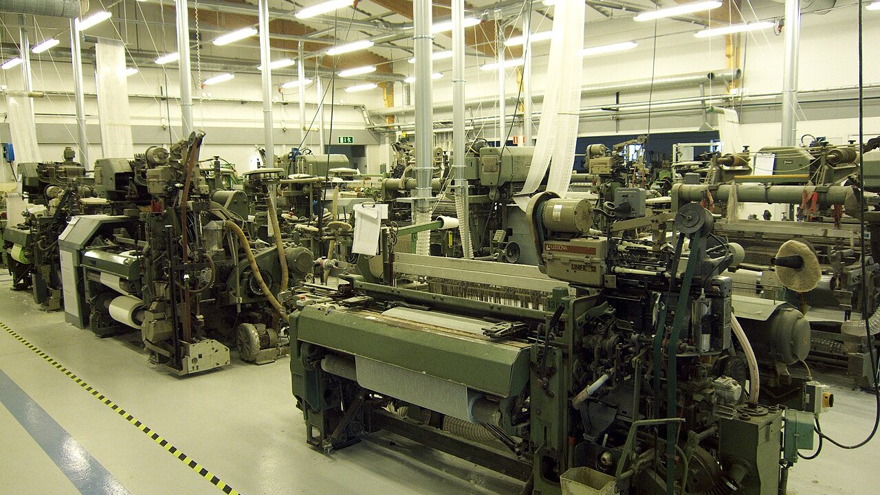A row of industrial weaving looms running in a bright factory hall.