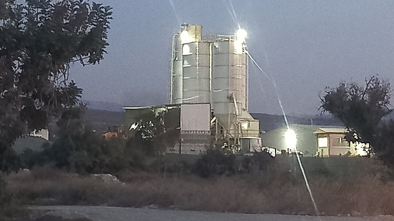 Tall grey cement silos and a processing building at dusk beside a dry field.