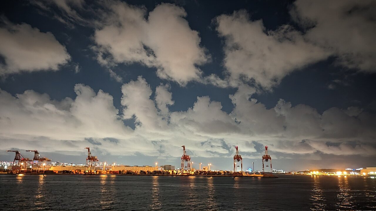 Container cranes and lit port terminal at night beside a calm harbour under cloudy skies