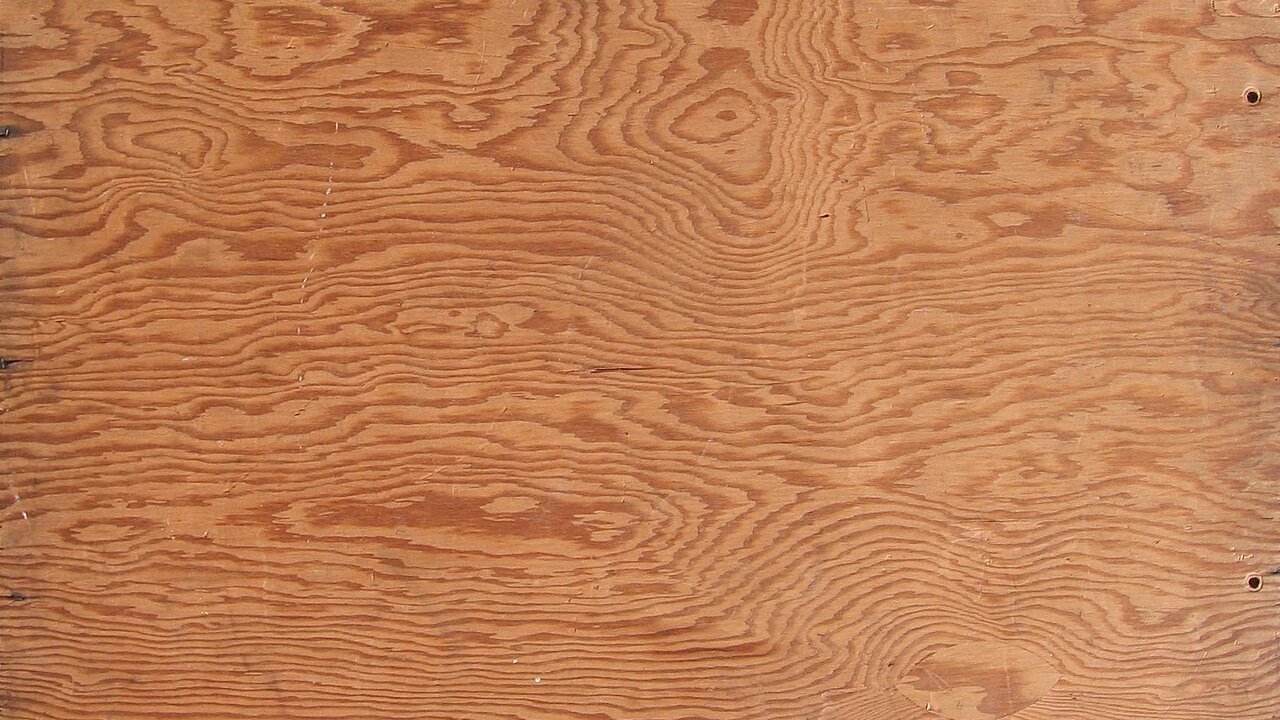 A close-up of a wood grain plywood sheet with swirling brown grain patterns.